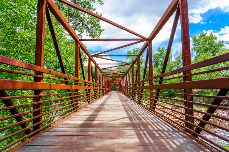 A pedestrian bridge over the Cache la Poudre River in Fort Collins, framed by lush trees under a bright sky